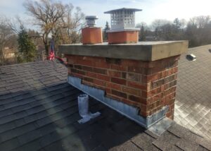 A brick chimney with two caps on a shingled roof, featuring an American flag in the background.